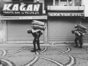 Black and white photo of two elderly men carrying heavy sacks on an urban street with closed shops.