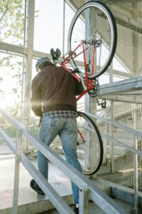 A man wearing a sweater ascends stairs carrying a bicycle, sunlit from large windows.
