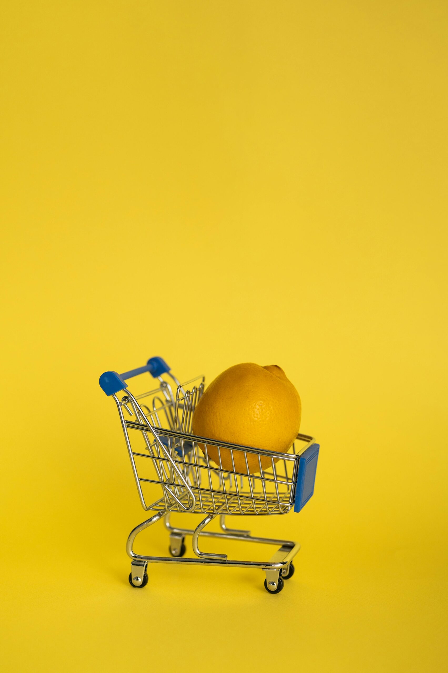 A single lemon sits inside a tiny shopping cart against a vibrant yellow background, creating a minimalist and bright composition.