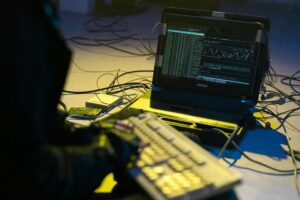A person typing on a keyboard connected to a laptop showing code, surrounded by wires.
